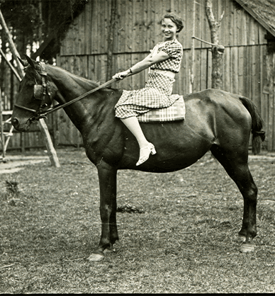 Młoda kobieta siedzi na koniu. Za nimi widoczny jest duży drewniany budynek. Kobieta ubrana jest w sukienkę w kratkę. Uśmiecha się. Link do podstrony z tym zdjęciem