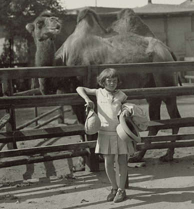 Dziewczynka stoi w ogrodzie zoologicznym oparta o drewniane ogrodzenie. Ubrana w sukienkę w dłoniach trzyma dwa kapelusze i płaszczyk. Za ogrodzeniem na wybiegu dla zwierząt stoi wielbłąd i spogląda w górę. Link do podstrony archiwum Stowarzyszenia Krotochwile